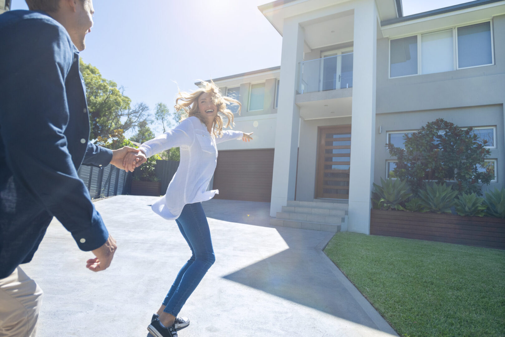 Couple joyfully running into their new home. They are both wearing casual clothes, very happy and smiling. The house is contemporary double brick with render, driveway and balcony. They are holding hands.