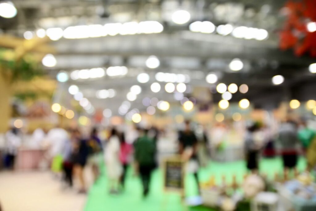 Blurred image of people in a brightly lit indoor event space with green flooring.